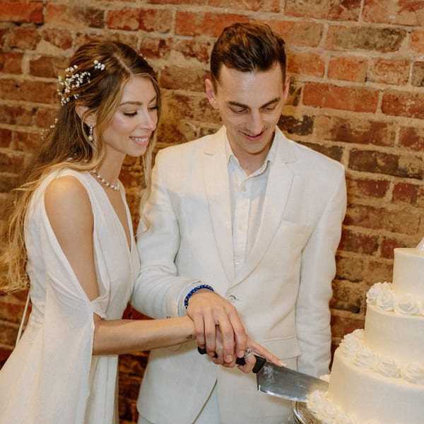 A smiling couple in white outfits cuts a white tiered cake with rose decorations against a brick wall.