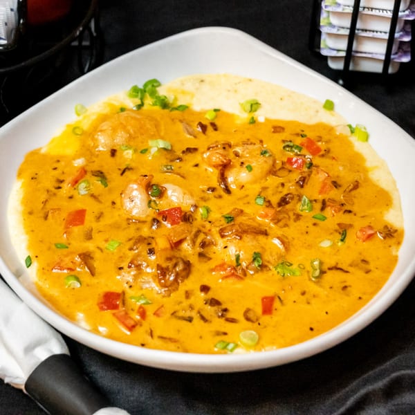 Shrimp and grits with a rich orange sauce, red peppers, and green onions in a white bowl.