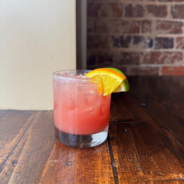 Pink cocktail with ice and an orange and lime wedge on a wooden table.