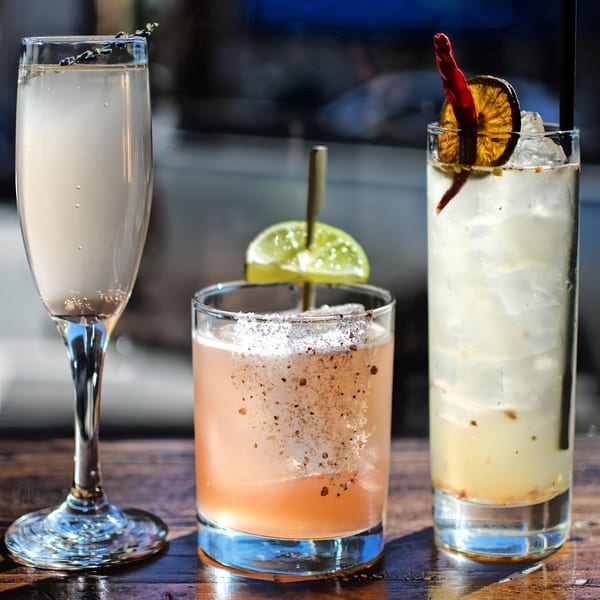 Three Honeycomb Cafe cocktails on a wooden table: a champagne flute, a pink drink with lime, and an iced drink with chili.
