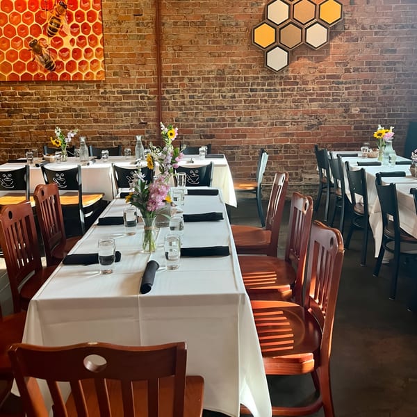Honeycomb Cafe dining room with white-clothed tables, wooden chairs, and bee-themed decor on a brick wall.