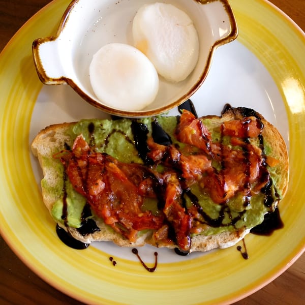Avocado toast with tomatoes and balsamic glaze, served with two poached eggs on a yellow plate.
