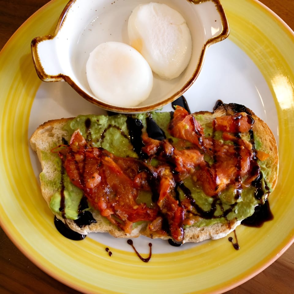 Avocado toast with tomatoes and balsamic glaze, served with two poached eggs on a yellow plate.