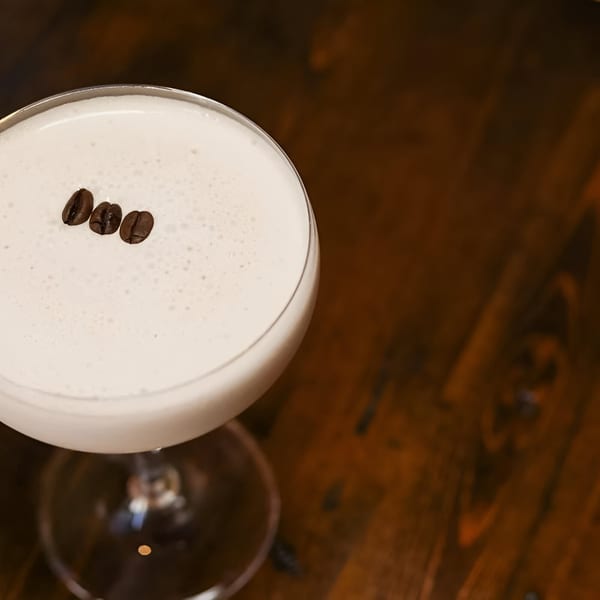 A creamy white cocktail in a martini glass, garnished with three coffee beans on a dark wooden surface.