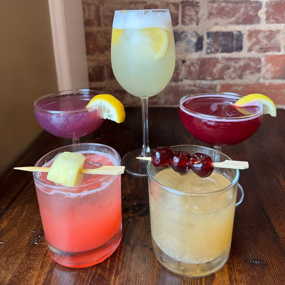 Four colorful Honeycomb Cafe cocktails, two with lemon slices, one with pineapple, and one with cherries.