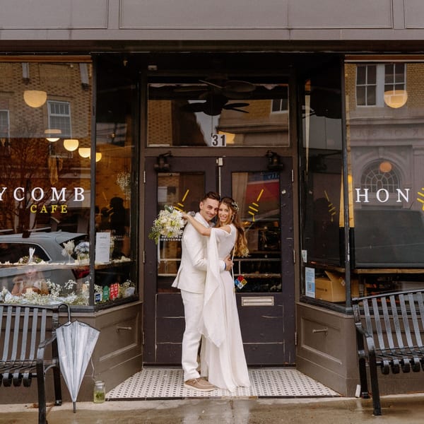 A newlywed couple in white embracing in front of the Honeycomb Cafe entrance.