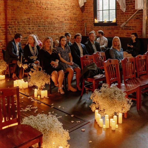 Guests seated in rows of wooden chairs, surrounded by candles and baby's breath, in a rustic room.