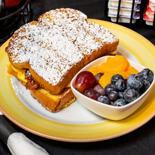 Powdered sugar French toast with bacon and scrambled eggs, served with a side of fresh fruit.